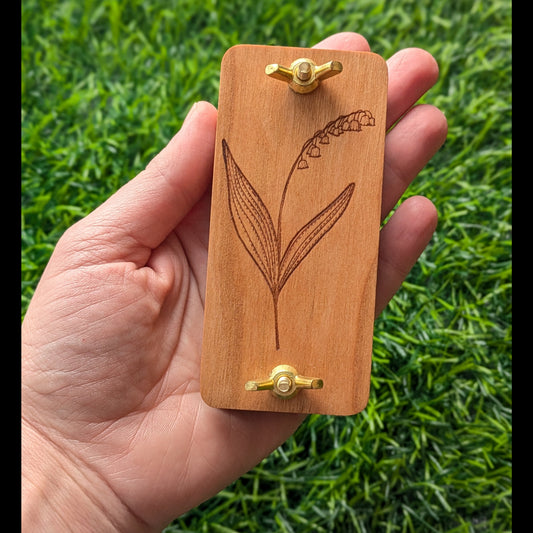Hand holding a wooden mini flower press with engraved Snowdrop flower design against a grass background