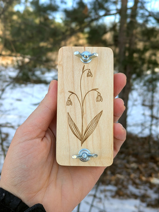 Hand holding a wooden mini flower press with engraved Snowdrop flower design against a blurred natural background